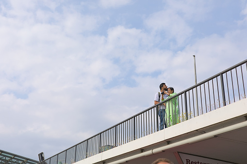 Hamburg, Germany - 26/08/2023: Arab couple in jeans and a green dress behind a railing on a bridge in front of a blue sky with white clouds