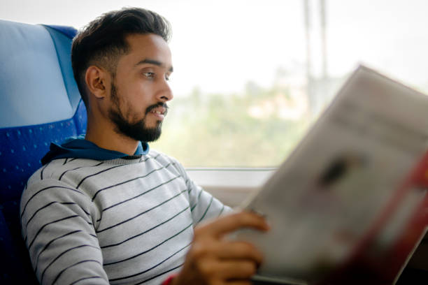 Portrait of an Indian young man sitting next to the window and reading a newspaper while traveling by the train.