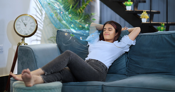 Happy gen z beautiful smiling woman sitting rest sofa raising arms stretching back muscles enjoy feel peaceful weekend at home. Indian gen z lazy female leaning relax on couch with closed eyes hands behind head spend time