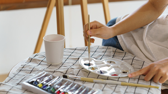 Young asian artist women using paintbrush to mixing color in color palette for painting picture in canvas on the easel while creative to drawing artwork with art hobby lifestyle in workshop studio.