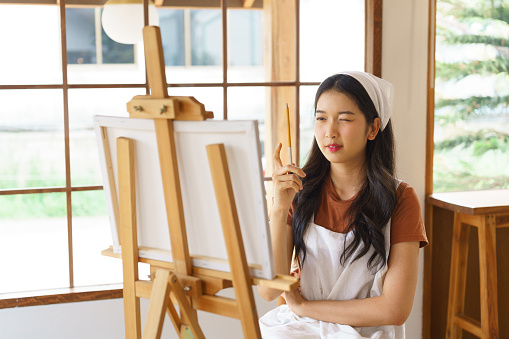 Young asian artist women holding paintbrush to proportion and painting masterpiece in canvas on the easel while creative to drawing artwork and working with art hobby lifestyle in workshop studio.