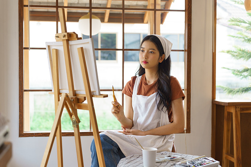 Young asian artist women holding paintbrush to proportion and painting masterpiece in canvas on the easel while creative to drawing artwork and working with art hobby lifestyle in workshop studio.