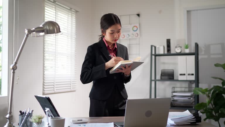 The focused young Asian businesswoman diligently records project details in her notebook at her office desk