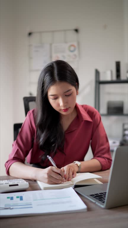 The focused young Asian businesswoman diligently records project details in her notebook at her office desk