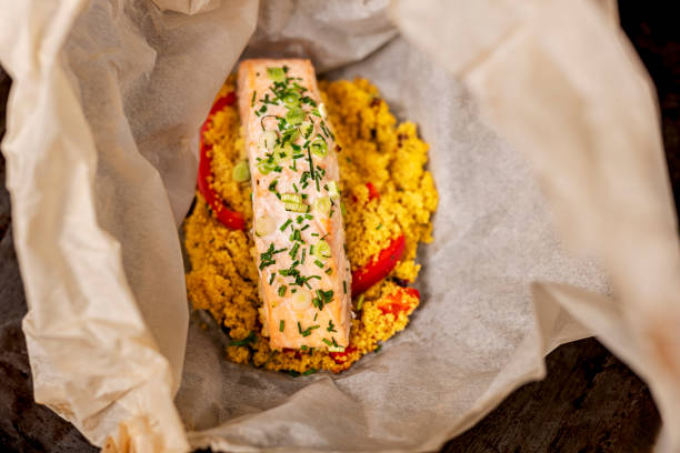 High-angle view of a dish of poached salmon with couscous. The whole dish is cooked together “en papillote”. Make your couscous according to the packet instructions place it on some oven-proof paper and put your salmon fillet on top, season it with some salt, pepper, chives and scallions then fold over the paper until you make a bag, and cook at 180C for around 15minutes depending on the size of your piece of salmon. Couscous is an alternative to pasta, it is healthy, low in fat, high in selenium and a good source of plant-based protein. The salmon is poached and then flaked and added to the couscous providing a rich source of omega 3 fatty acids  and vitamin B12.