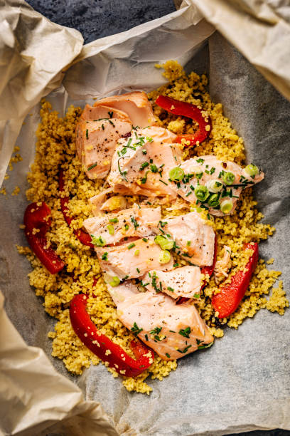 High-angle, view of a dish of poached salmon with couscous. The whole dish is cooked together “en papillote”. Make your couscous according to the packet instructions place it on some oven-proof paper and put your salmon fillet on top, season it with some salt, pepper, chives and scallions then fold over the paper until you make a bag, and cook at 180C for around 15minutes depending on the size of your piece of salmon. Couscous is an alternative to pasta, it is healthy, low in fat, high in selenium and a good source of plant-based protein. The salmon is poached and then flaked and added to the couscous providing a rich source of omega 3 fatty acids  and vitamin B12.