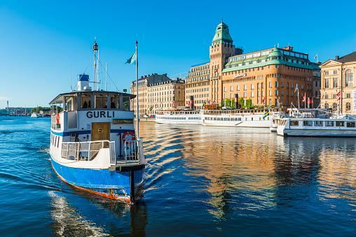 Warm summer sunlight illuminating the ferries on the harbour waterfront reflecting the iconic facades of the luxury hotels and townhouses that line the shore of Nybroviken harbour, Stockholm, Sweden's vibrant capital city.