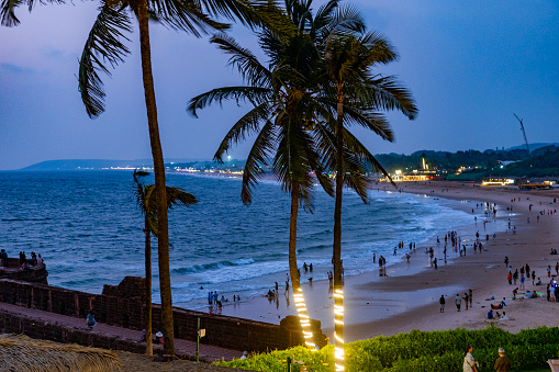 A beach at sunset with tall palm trees, people walking along the shore in Hikkaduwa. The waves are rolling in under a sky with soft clouds illuminated by the setting sun. The tall palm trees stand out against the backdrop of a cloudy yet bright sky. People are seen walking along the shore, enjoying the view and atmosphere.  The ocean waves roll towards the sandy shore under an illuminated sky. The clouds in the sky are soft and diffused by sunlight.