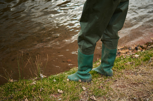 Fisherman boots in green color standing by the river on muddy ground grass