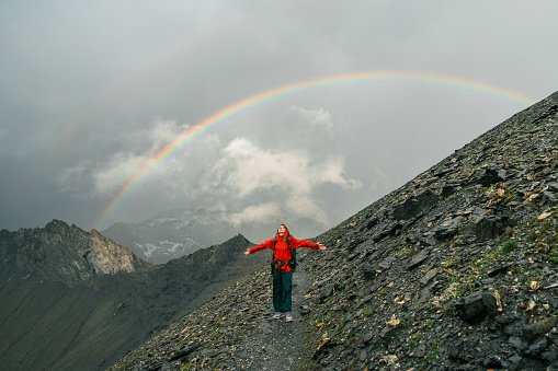 Woman with arms raised looks with awe at the landscape during the hike under the rain in Swiss Alps. She is standing under the rainbow