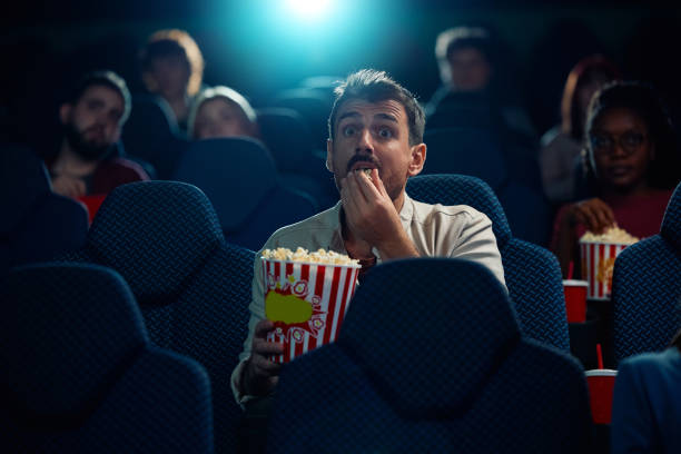 Young man watching suspenseful film projection in movie theater. Young man eating popcorn while watching movie in disbelief in cinema. suspenseful stock pictures, royalty-free photos & images