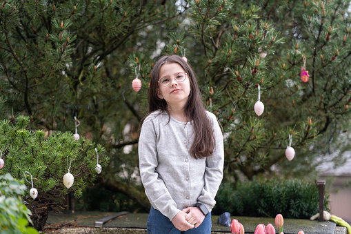 Cute little girl with long brown hair and freckles in a gray sweater stands near a tree with Easter eggs