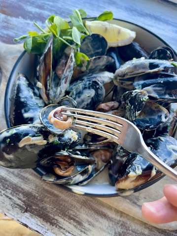 High angle closeup photo of a bowl of cooked Australian black mussels with a lemon wedge and pea shoots garnish. A woman’s fingers hold a fork with a mussel.