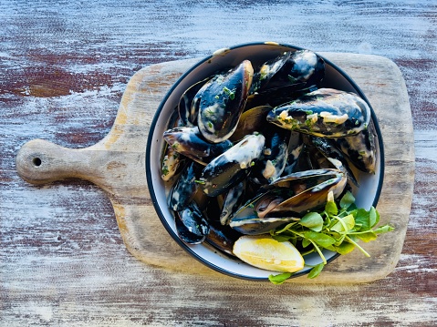 High angle closeup photo of a bowl of cooked Australian black mussels with a white wine and cream sauce and a lemon wedge and pea shoot garnish, served on a wooden board on a rustic recycled wooden bench in a fine dining restaurant in Summer.