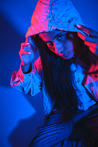 A woman with long hair is wearing a hooded jacket and sitting in front of a blue wall. The image has a moody and mysterious feel to it, as the woman's gaze is directed towards the camera