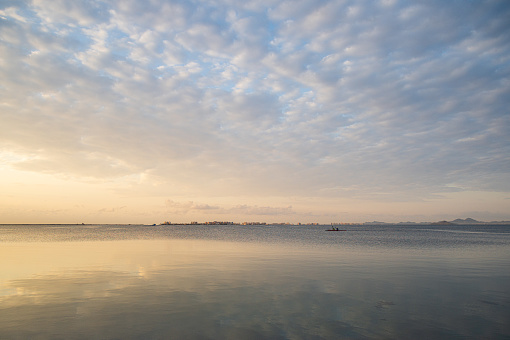 Calm waters of the Mar Menor in a stunning sunrise, with a canoe plying its waters. The morning clouds are reflected in the water and in the background you can see the town of La Manga del Mar Menor. An image that invites relaxation. Copy space