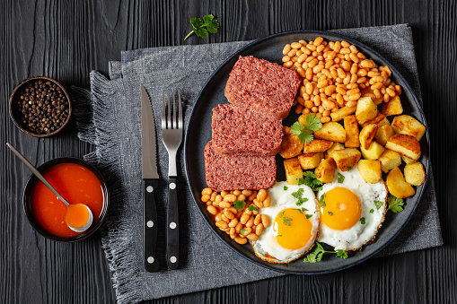 corned beef slices, baked beans, roasted potatoes and sunny side up fried eggs on plate on black wooden table with cutlery and hot sauce, horizontal view from above, flat lay
