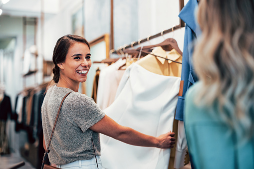 A cheerful young woman is selecting garments in a boutique store, sharing a joyous smile while browsing through racks of stylish clothing with a friend.