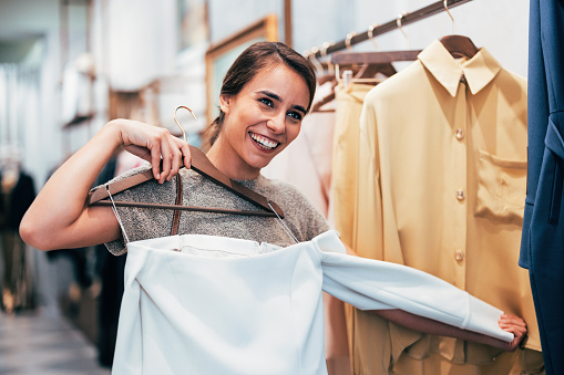 A cheerful young woman enjoys her time while picking out clothes in a boutique. She holds hangers with stylish garments, smiling brightly.