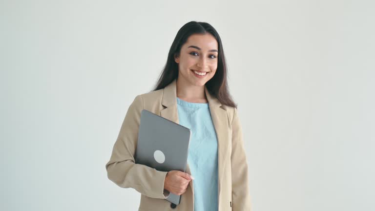 Female successful entrepreneur. Confident indian or arabian business woman, office worker in elegant suit, holding laptop, standing on isolated white background, looks at camera, smiling
