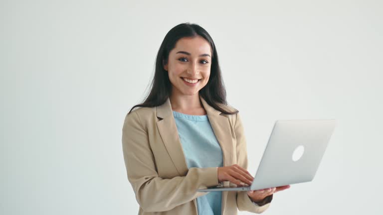 Business concept. Beautiful confident indian or arabian business woman, office worker in elegant suit, holding an open laptop, standing on isolated white background, looks at camera, smiling friendly