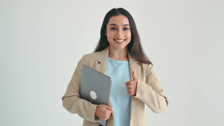 Confident indian or arabian business woman, office worker in elegant suit, holding laptop, standing on isolated white background, looks at camera, smiling, shows thumb-up gesture, agreement sign
