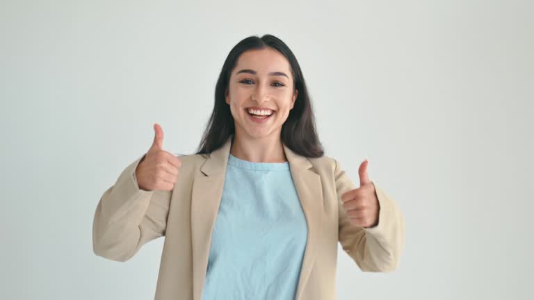 Happy indian or arabian brunette business woman, dressed in elegant suit, manager, gladly looks at camera and showing thumb-up gesture with both hands, standing on isolated white background, smile