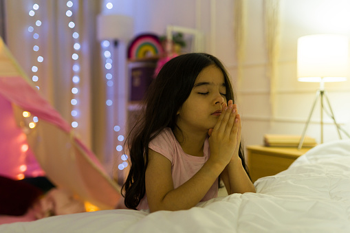 Young hispanic girl closes her eyes and clasps her hands in prayer in her cozy, warmly lit bedroom at night