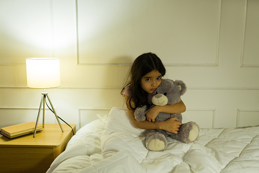 Hispanic little girl hugs her teddy bear tightly in the soft glow of her bedroom lamp during nighttime and looks affraid of nightmares