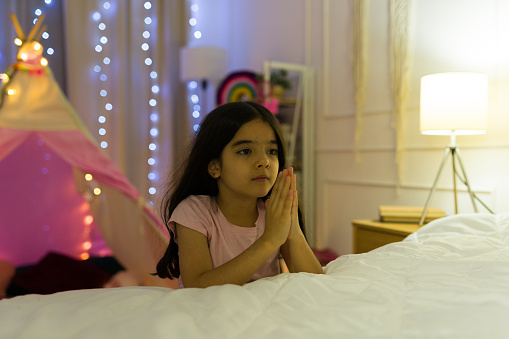 Young latin girl with a hopeful expression prays in her cozy, softly lit bedroom at home during the nighttime