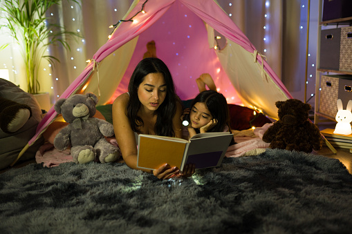 Mother and daughter enjoy reading together in a homemade tent with fairy lights, creating a warm atmosphere at home at night