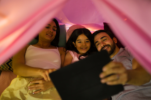 Happy family having a cozy evening, watching a movie inside a vibrant blanket fort with gentle lighting setting a cozy atmosphere