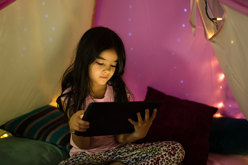 Young hispanic girl concentrates on her tablet inside a whimsically lit home bedroom, creating a magical nighttime atmosphere