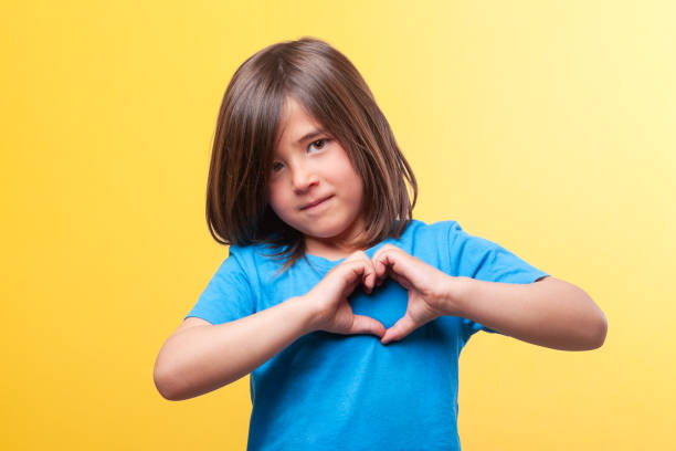 Boy with blue t-shirt and long brown hair makes the shape of a heart with his hands next to his chest. Boy with blue t-shirt and long brown hair makes the shape of a heart with his hands next to his chest while looking at the camera. cute boy show love symbol hand stock pictures, royalty-free photos & images