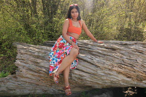 A Bangladeshi woman sitting on a weathered log. She is wearing medium length black hair, straight hair, makeup, earrings, an orange crop top, multi colored, floral print skirt and sandals.