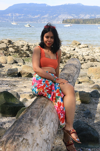 A Bangladeshi woman sitting on a log that has been washed up on a beach near Vancouver, Canada. She is wearing medium length black hair, sunglasses, makeup, and orange crop top, a multicolored floral print dress, red open toe shoes and bracelets.