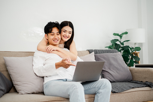 Young Asian couple enjoys a cozy moment on their sofa, interacting with a laptop, in a light-filled, plant-adorned living space.
