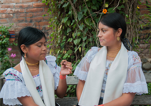 Two young indigenous girls have a conversation while sharing a piece of chocolate, dressed in their traditional costumes, against the backdrop of a brick wall intertwined with green foliage. High quality photo