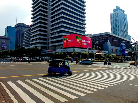 A Jakarta cityscape view of the Bajaj, three-wheeled vehicle or rickshaw, drives and passes through the Sarinah pelican crossing on the road of M.H Thamrin in the morning.