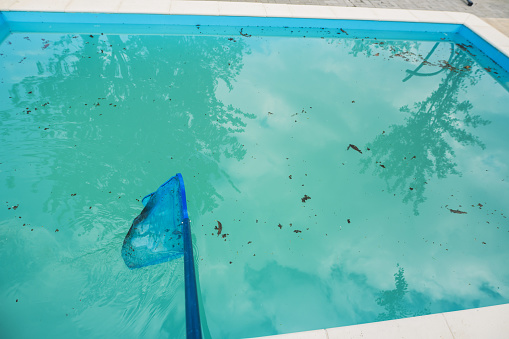 Stock photo showing close-up, elevated view of public swimming pool with turquoise blue mosaic tiles. A cracked, square slab on the surrounding patio area has caused a puddle do to a leak.