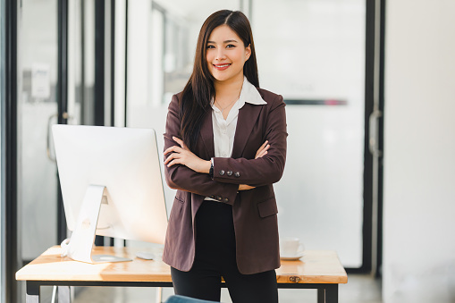 A friendly Asian businesswoman with folded arms smiles warmly, standing in front of her workspace in a modern office environment.