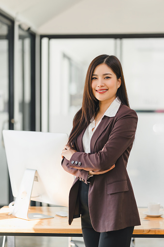 A friendly Asian businesswoman with folded arms smiles warmly, standing in front of her workspace in a modern office environment.