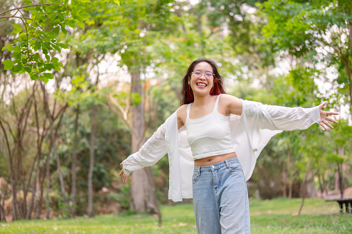 A cheerful, carefree young Asian woman in glasses, wearing a white top, and denim jeans, smiles warmly while spinning around in a green park, showing a joyful expression.