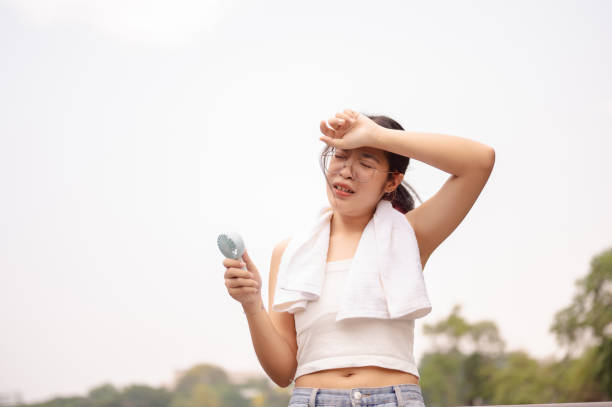 A tired woman holds a small fan and wipes her sweat on her face while walking outdoors on a hot day.