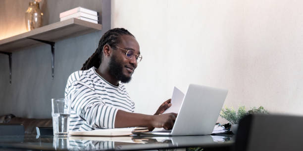 istock African American man working with laptop computer remote while sitting at glass table in living room. Black guy do freelance work at home office 2150229932