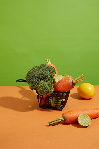 Green background against on black basket contained some vegetable including broccoli, carrot and lime on orange kitchen counter. Front view with space for advertising