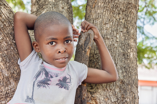 small village african girl, standing in in the yard , arms up in front of a tree trunk
