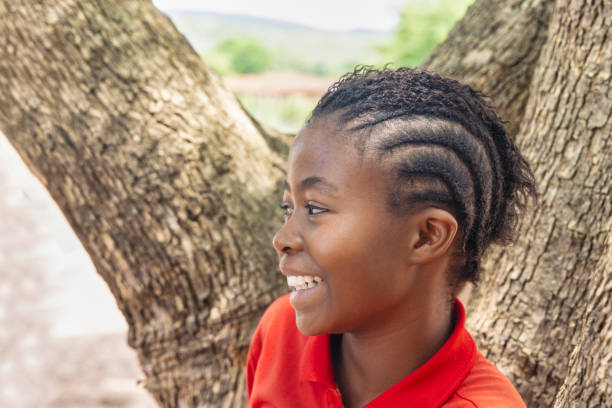 village african teenager girl profile view , wearing braids hairstyle , standing in front of a tree in the yard