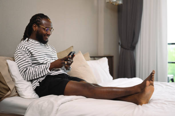 istock Smiling African guy checking messages on his mobile phone lying in bed at home 2150236614
