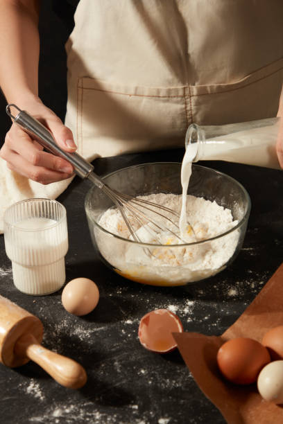istock The baker is pouring milk from the bottle into a bowl containing flour and eggs, and using a whisk to mix it. A cup filled with milk and a rolling pin displayed on black table with eggs 2150237278